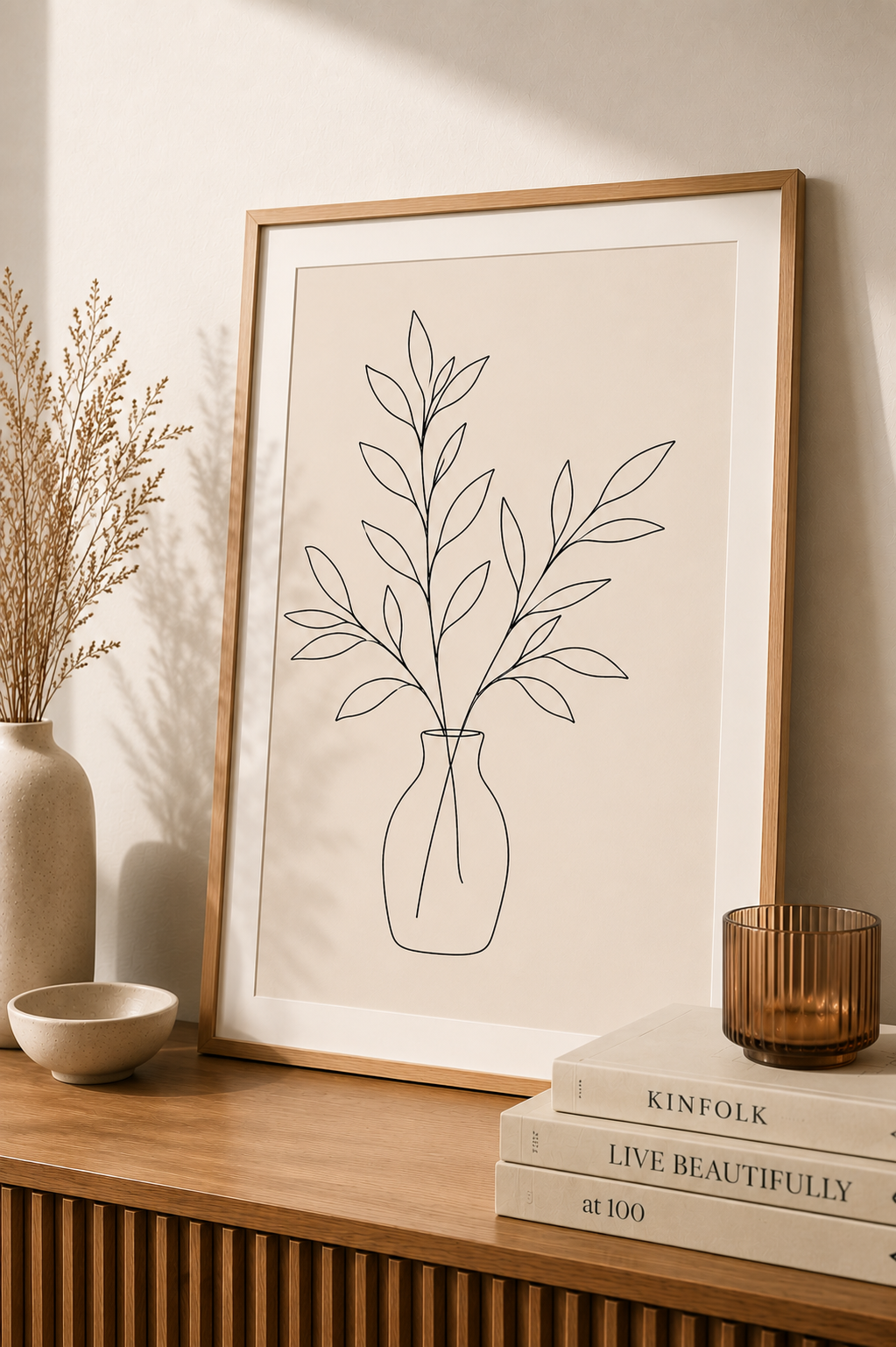 Botanical Vase minimal line art print — framed wall art mockup leaning on a warm beige wall above a wooden sideboard with books and candle, Morphē Studio