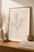 Botanical Vase minimal line art print — framed wall art mockup leaning on a warm beige wall above a wooden sideboard with books and candle, Morphē Studio