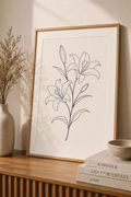 Lilly Bloom minimal line art print — framed botanical wall art leaning on a warm beige wall above a wooden sideboard with dried pampas grass, Kinfolk books and ceramic bowl, Morphē Studio