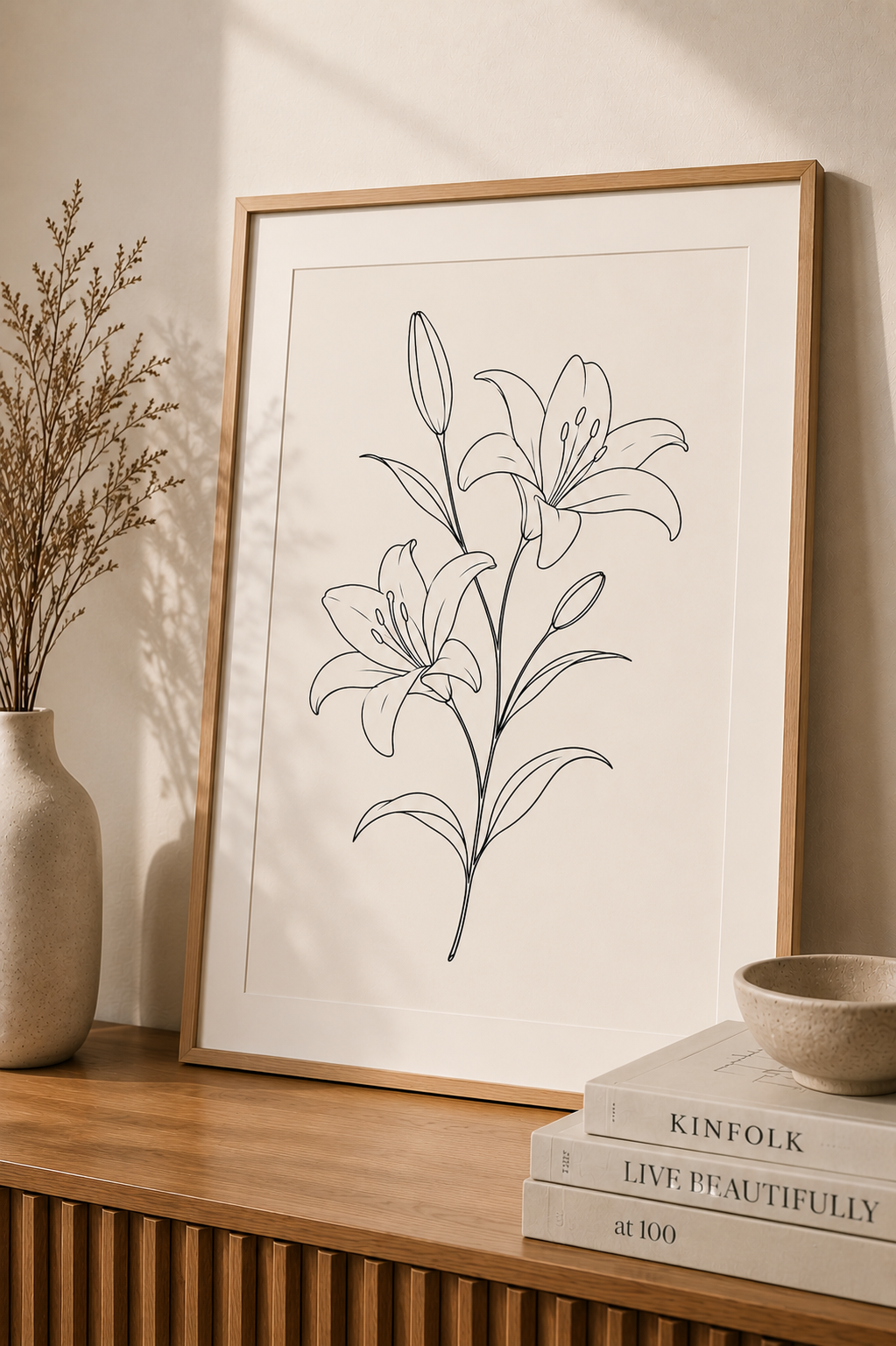 Lilly Bloom minimal line art print — framed botanical wall art leaning on a warm beige wall above a wooden sideboard with dried pampas grass, Kinfolk books and ceramic bowl, Morphē Studio