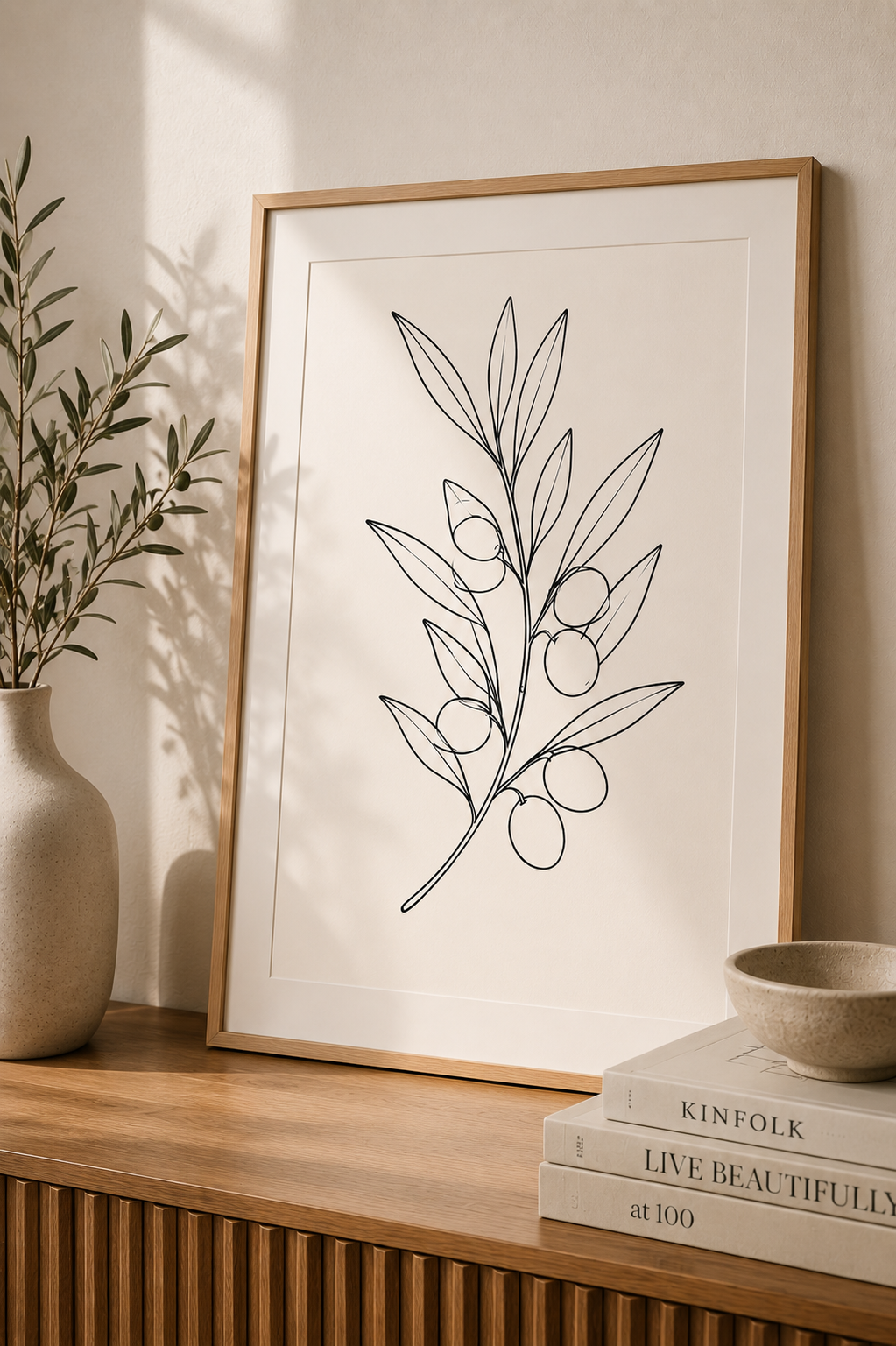 Olive Branch minimal line art print — framed botanical wall art leaning on a warm beige wall above a wooden sideboard with olive branch vase, Kinfolk books and ceramic bowl, Morphē Studio