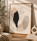 Quiet Embrace minimal line art print — large framed wall art of a couple in an intimate embrace with taupe shadow accents, leaning on a warm cream wall in a Scandinavian living room with olive tree and ceramic vases, Morphē Studio