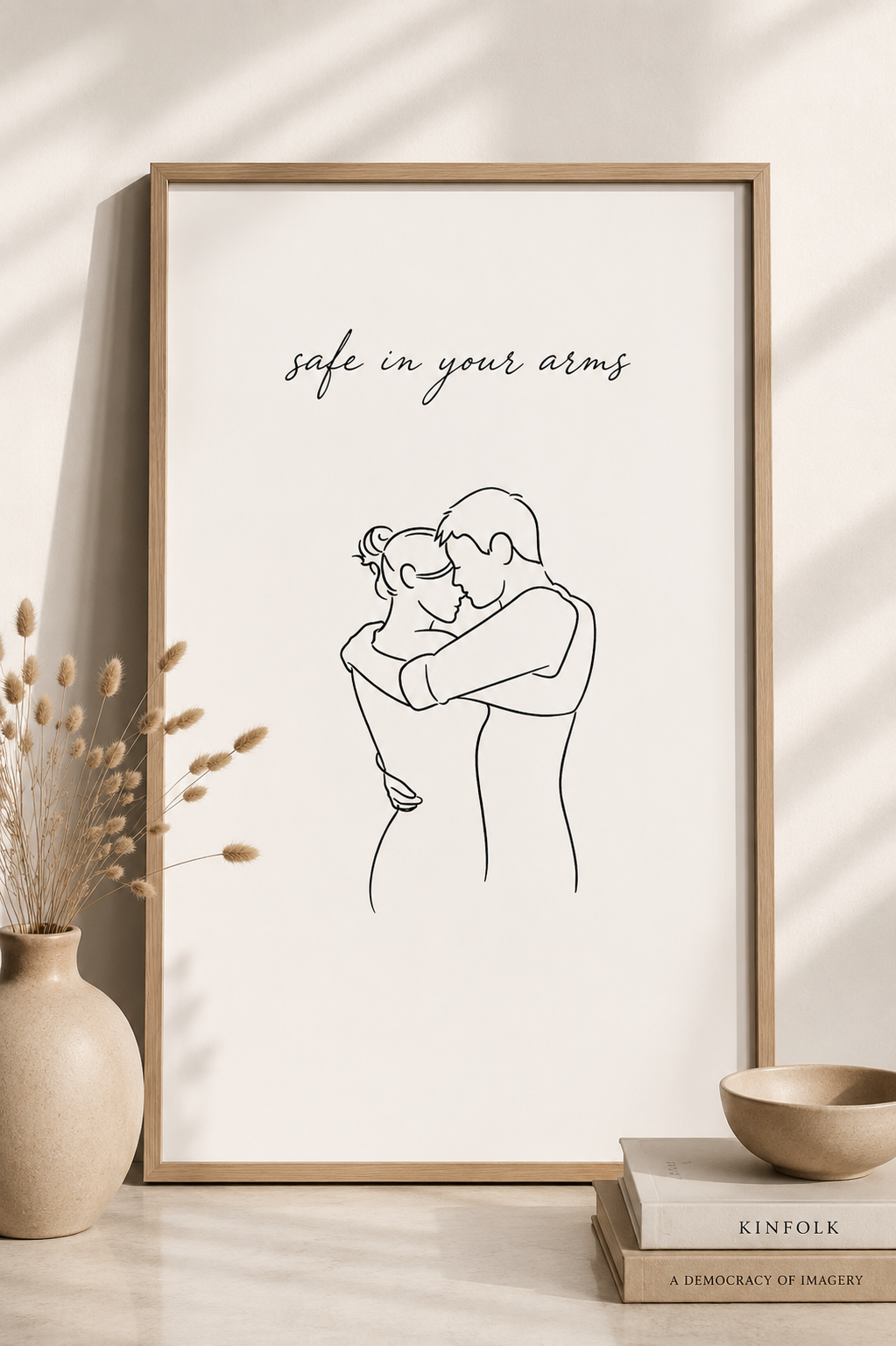 Safe in Your Arms minimal line art print — framed wall art with couple embrace and script typography, leaning on a warm white wall with dried pampas grass vase, Kinfolk books and ceramic bowl, Morphē Studio