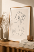 Soft Back Muse minimal line art print — framed wall art of a woman from behind with elegant updo and draped fabric, leaning on a warm beige wall above a wooden sideboard with dried grass, Kinfolk books and amber glass, Morphē Studio