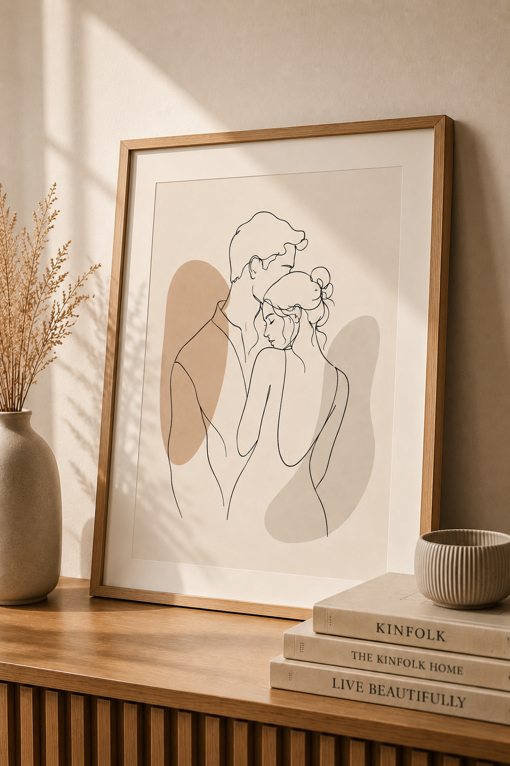 Tender Silence minimal line art print — framed wall art of a couple in a gentle embrace with taupe shadow accents, leaning on a warm beige wall above a wooden sideboard with dried pampas grass, Kinfolk books and ribbed ceramic bowl, Morphē Studio