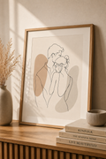 Tender Silence minimal line art print — framed wall art of a couple in a gentle embrace with taupe shadow accents, leaning on a warm beige wall above a wooden sideboard with dried pampas grass, Kinfolk books and ribbed ceramic bowl, Morphē Studio