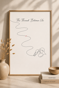The Thread Between Us minimal line art print — large framed wall art with red thread of fate winding down to a couple facing each other, leaning on a warm beige wall with dried pampas grass vase, Kinfolk books and ceramic bowl, Morphē Studio